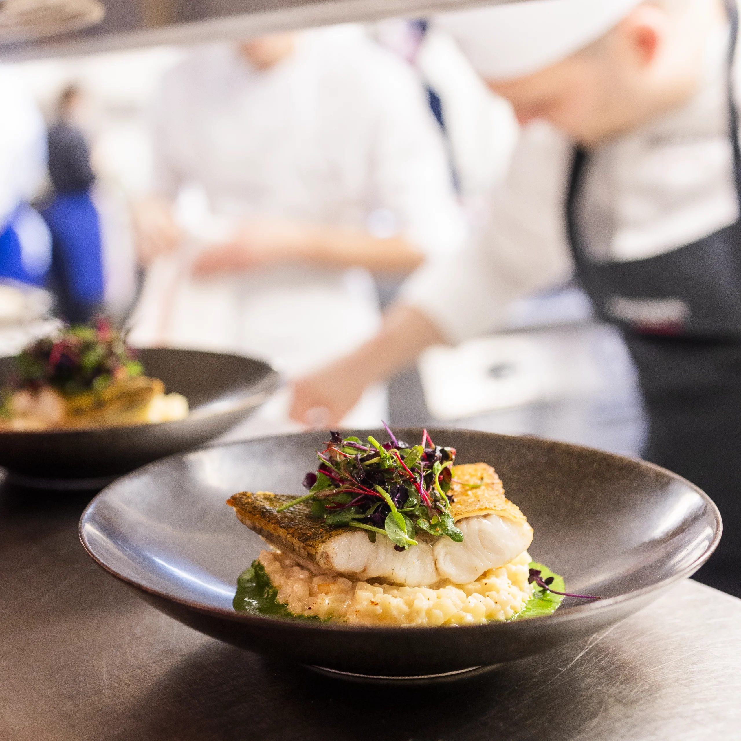 plat gastronomique à base de poisson dans une assiette noire avec un cuisinier en arrière plan